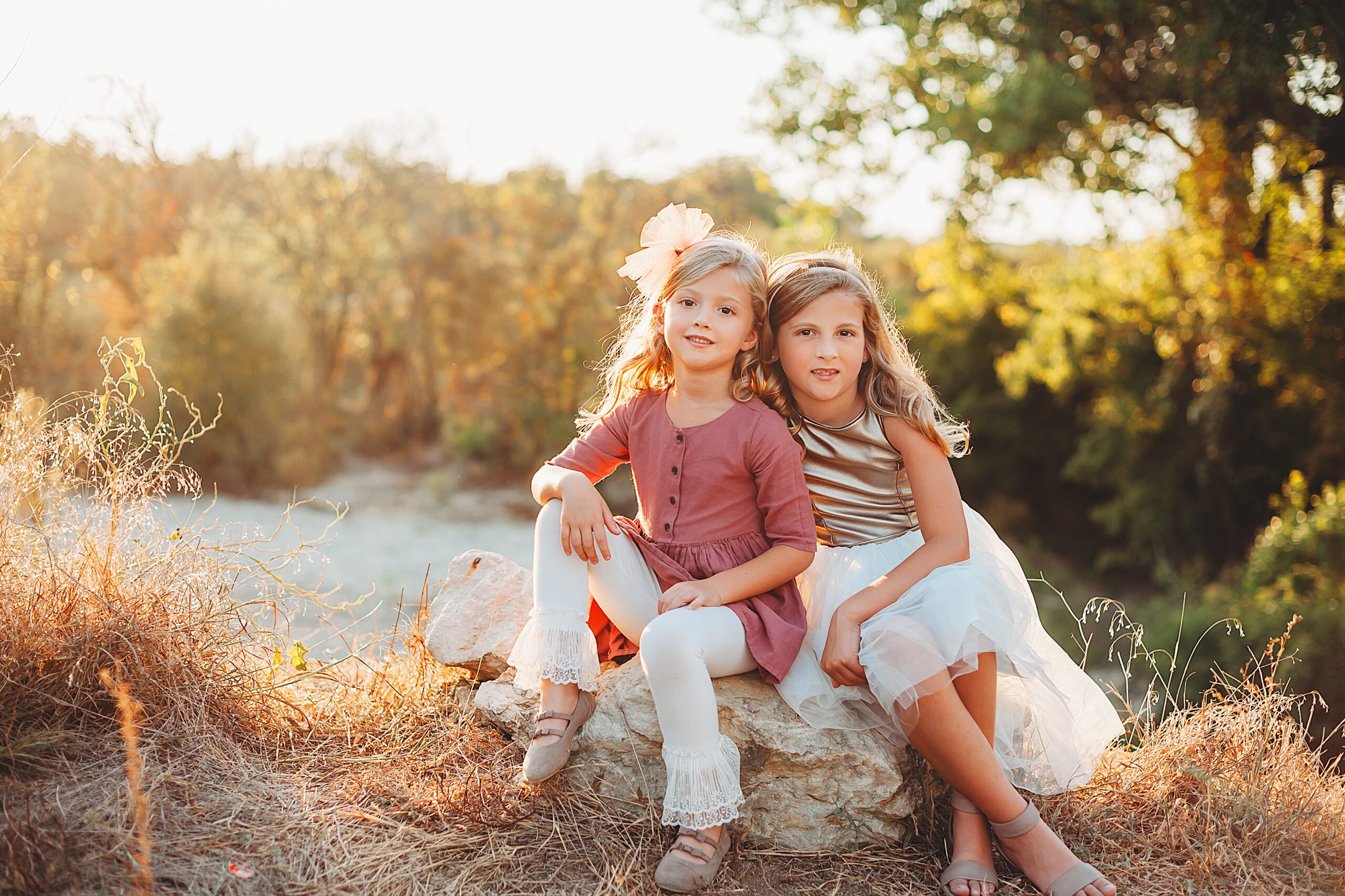 Two sisters sit smiling in the golden sunshine in a Fort Worth Family Photography session by Splendor Photography